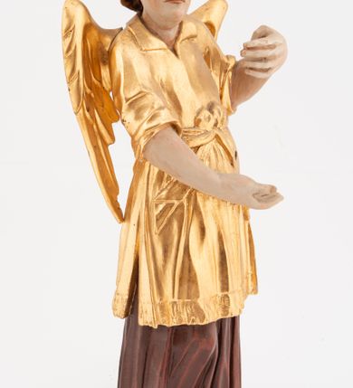 Zdjęcie nr 3: Figura pełnoplastyczna, ustawiona na niskiej podstawie. Postać ukazana frontalnie, z głową skierowaną w prawo, w kontrapoście, z obiema rękami uniesionymi z przodu, jedną wysoko, drugą nisko. Twarz ma podłużną, z drugim podbródkiem, o dużych, migdałowatych i opadających oczach, wąskim nosie i ustach, okoloną krótkimi, kręconymi włosami, układającymi się w czub nad czołem. Anioł ubrany jest w ciemnoczerwoną suknię spodnią i złoconą suknię wierzchnią, sięgającą kolan, z podwiniętymi rękawami, przepasaną w talii szarfą. Skrzydła ma złocone, złożone na plecach. Polichromia ciała naturalistyczna. 