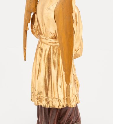 Zdjęcie nr 5: Figura pełnoplastyczna, ustawiona na niskiej podstawie. Postać ukazana frontalnie, z głową skierowaną w prawo, w kontrapoście, z obiema rękami uniesionymi z przodu, jedną wysoko, drugą nisko. Twarz ma podłużną, z drugim podbródkiem, o dużych, migdałowatych i opadających oczach, wąskim nosie i ustach, okoloną krótkimi, kręconymi włosami, układającymi się w czub nad czołem. Anioł ubrany jest w ciemnoczerwoną suknię spodnią i złoconą suknię wierzchnią, sięgającą kolan, z podwiniętymi rękawami, przepasaną w talii szarfą. Skrzydła ma złocone, złożone na plecach. Polichromia ciała naturalistyczna. 