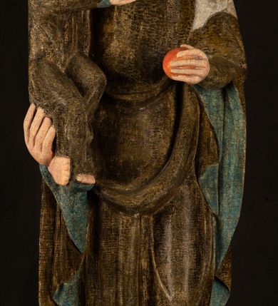 Zdjęcie nr 13: Stojąca figura Matki Boskiej z Dzieciątkiem reprezentuje przykład przyściennej rzeźby z tyłu wydrążonej. Maria stoi w lekkim kontrapoście, podkreślonym wysunięciem do przodu lewego kolana na niewielkim wzgórku. Prawą ręką podtrzymuje Dzieciątko, w lewej dłoni trzyma jabłko. Madonna ma mocno pociągłą twarz o jasnej karnacji z wysokim czołem, rumianych policzkach, długi, prosty nos i wydatne usta. Jej głowa osadzona jest na długiej szyi. Spod białej chusty ubranej na głowę i opadającej na ramiona widać brązowe włosy. Matka Boska na głowie ma koronę o ostrych, krótkich sterczynach. Nałożony na ciemnooliwkową suknię płaszcz w tym samym kolorze, podbity błękitem, tworzy dwa wałkowate zaokrąglone fałdy na brzuchu Marii, spływając miękkimi fałdami spod lewej ręki oraz festonem spod prawej ręki. Niżej fałdy płaszcza i sukni układają się niemal pionowo, opinając materią lewą nogę. Długa suknia odsłania czubki ciżemek. Dzieciątko zostało ujęte w trzech czwartych w prawo. Obie jego rączki wyciągnięte są przed siebie w stronę trzymanego przez Marię jabłka. Z kolei nóżki są skrzyżowane przy stopach. Jezus ma wyrazistą twarz o jasnej karnacji, małym nosem i wydatnych ustach. Jego głowę otaczają brązowe włosy układające się po bokach w charakterystyczne pukle. Jezus ubrany jest w długą, ciemnooliwkową tunikę odsłaniającą fragmenty stóp z charakterystycznymi mankietami przy rękawach. Szaty Matki Boskiej i Dzieciątka są srebrzone i laserowane.