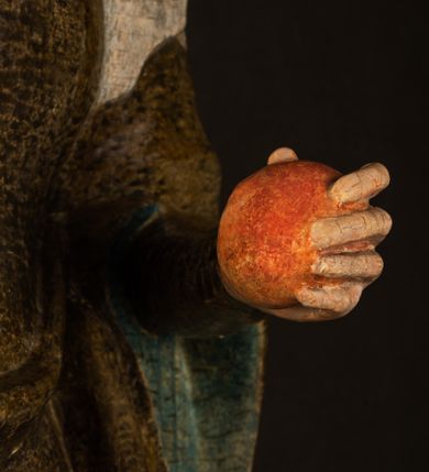 Zdjęcie nr 16: Stojąca figura Matki Boskiej z Dzieciątkiem reprezentuje przykład przyściennej rzeźby z tyłu wydrążonej. Maria stoi w lekkim kontrapoście, podkreślonym wysunięciem do przodu lewego kolana na niewielkim wzgórku. Prawą ręką podtrzymuje Dzieciątko, w lewej dłoni trzyma jabłko. Madonna ma mocno pociągłą twarz o jasnej karnacji z wysokim czołem, rumianych policzkach, długi, prosty nos i wydatne usta. Jej głowa osadzona jest na długiej szyi. Spod białej chusty ubranej na głowę i opadającej na ramiona widać brązowe włosy. Matka Boska na głowie ma koronę o ostrych, krótkich sterczynach. Nałożony na ciemnooliwkową suknię płaszcz w tym samym kolorze, podbity błękitem, tworzy dwa wałkowate zaokrąglone fałdy na brzuchu Marii, spływając miękkimi fałdami spod lewej ręki oraz festonem spod prawej ręki. Niżej fałdy płaszcza i sukni układają się niemal pionowo, opinając materią lewą nogę. Długa suknia odsłania czubki ciżemek. Dzieciątko zostało ujęte w trzech czwartych w prawo. Obie jego rączki wyciągnięte są przed siebie w stronę trzymanego przez Marię jabłka. Z kolei nóżki są skrzyżowane przy stopach. Jezus ma wyrazistą twarz o jasnej karnacji, małym nosem i wydatnych ustach. Jego głowę otaczają brązowe włosy układające się po bokach w charakterystyczne pukle. Jezus ubrany jest w długą, ciemnooliwkową tunikę odsłaniającą fragmenty stóp z charakterystycznymi mankietami przy rękawach. Szaty Matki Boskiej i Dzieciątka są srebrzone i laserowane.