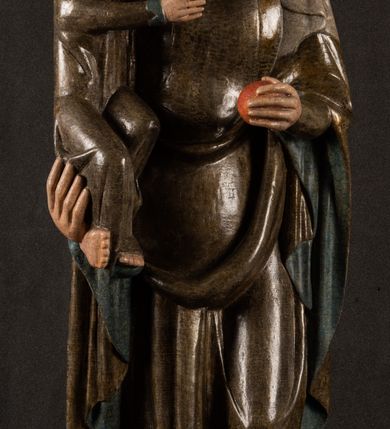 Zdjęcie nr 22: Stojąca figura Matki Boskiej z Dzieciątkiem reprezentuje przykład przyściennej rzeźby z tyłu wydrążonej. Maria stoi w lekkim kontrapoście, podkreślonym wysunięciem do przodu lewego kolana na niewielkim wzgórku. Prawą ręką podtrzymuje Dzieciątko, w lewej dłoni trzyma jabłko. Madonna ma mocno pociągłą twarz o jasnej karnacji z wysokim czołem, rumianych policzkach, długi, prosty nos i wydatne usta. Jej głowa osadzona jest na długiej szyi. Spod białej chusty ubranej na głowę i opadającej na ramiona widać brązowe włosy. Matka Boska na głowie ma koronę o ostrych, krótkich sterczynach. Nałożony na ciemnooliwkową suknię płaszcz w tym samym kolorze, podbity błękitem, tworzy dwa wałkowate zaokrąglone fałdy na brzuchu Marii, spływając miękkimi fałdami spod lewej ręki oraz festonem spod prawej ręki. Niżej fałdy płaszcza i sukni układają się niemal pionowo, opinając materią lewą nogę. Długa suknia odsłania czubki ciżemek. Dzieciątko zostało ujęte w trzech czwartych w prawo. Obie jego rączki wyciągnięte są przed siebie w stronę trzymanego przez Marię jabłka. Z kolei nóżki są skrzyżowane przy stopach. Jezus ma wyrazistą twarz o jasnej karnacji, małym nosem i wydatnych ustach. Jego głowę otaczają brązowe włosy układające się po bokach w charakterystyczne pukle. Jezus ubrany jest w długą, ciemnooliwkową tunikę odsłaniającą fragmenty stóp z charakterystycznymi mankietami przy rękawach. Szaty Matki Boskiej i Dzieciątka są srebrzone i laserowane.
