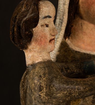 Zdjęcie nr 20: Stojąca figura Matki Boskiej z Dzieciątkiem reprezentuje przykład przyściennej rzeźby z tyłu wydrążonej. Maria stoi w lekkim kontrapoście, podkreślonym wysunięciem do przodu lewego kolana na niewielkim wzgórku. Prawą ręką podtrzymuje Dzieciątko, w lewej dłoni trzyma jabłko. Madonna ma mocno pociągłą twarz o jasnej karnacji z wysokim czołem, rumianych policzkach, długi, prosty nos i wydatne usta. Jej głowa osadzona jest na długiej szyi. Spod białej chusty ubranej na głowę i opadającej na ramiona widać brązowe włosy. Matka Boska na głowie ma koronę o ostrych, krótkich sterczynach. Nałożony na ciemnooliwkową suknię płaszcz w tym samym kolorze, podbity błękitem, tworzy dwa wałkowate zaokrąglone fałdy na brzuchu Marii, spływając miękkimi fałdami spod lewej ręki oraz festonem spod prawej ręki. Niżej fałdy płaszcza i sukni układają się niemal pionowo, opinając materią lewą nogę. Długa suknia odsłania czubki ciżemek. Dzieciątko zostało ujęte w trzech czwartych w prawo. Obie jego rączki wyciągnięte są przed siebie w stronę trzymanego przez Marię jabłka. Z kolei nóżki są skrzyżowane przy stopach. Jezus ma wyrazistą twarz o jasnej karnacji, małym nosem i wydatnych ustach. Jego głowę otaczają brązowe włosy układające się po bokach w charakterystyczne pukle. Jezus ubrany jest w długą, ciemnooliwkową tunikę odsłaniającą fragmenty stóp z charakterystycznymi mankietami przy rękawach. Szaty Matki Boskiej i Dzieciątka są srebrzone i laserowane.