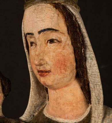 Zdjęcie nr 19: Stojąca figura Matki Boskiej z Dzieciątkiem reprezentuje przykład przyściennej rzeźby z tyłu wydrążonej. Maria stoi w lekkim kontrapoście, podkreślonym wysunięciem do przodu lewego kolana na niewielkim wzgórku. Prawą ręką podtrzymuje Dzieciątko, w lewej dłoni trzyma jabłko. Madonna ma mocno pociągłą twarz o jasnej karnacji z wysokim czołem, rumianych policzkach, długi, prosty nos i wydatne usta. Jej głowa osadzona jest na długiej szyi. Spod białej chusty ubranej na głowę i opadającej na ramiona widać brązowe włosy. Matka Boska na głowie ma koronę o ostrych, krótkich sterczynach. Nałożony na ciemnooliwkową suknię płaszcz w tym samym kolorze, podbity błękitem, tworzy dwa wałkowate zaokrąglone fałdy na brzuchu Marii, spływając miękkimi fałdami spod lewej ręki oraz festonem spod prawej ręki. Niżej fałdy płaszcza i sukni układają się niemal pionowo, opinając materią lewą nogę. Długa suknia odsłania czubki ciżemek. Dzieciątko zostało ujęte w trzech czwartych w prawo. Obie jego rączki wyciągnięte są przed siebie w stronę trzymanego przez Marię jabłka. Z kolei nóżki są skrzyżowane przy stopach. Jezus ma wyrazistą twarz o jasnej karnacji, małym nosem i wydatnych ustach. Jego głowę otaczają brązowe włosy układające się po bokach w charakterystyczne pukle. Jezus ubrany jest w długą, ciemnooliwkową tunikę odsłaniającą fragmenty stóp z charakterystycznymi mankietami przy rękawach. Szaty Matki Boskiej i Dzieciątka są srebrzone i laserowane.