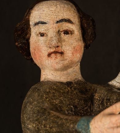 Zdjęcie nr 21: Stojąca figura Matki Boskiej z Dzieciątkiem reprezentuje przykład przyściennej rzeźby z tyłu wydrążonej. Maria stoi w lekkim kontrapoście, podkreślonym wysunięciem do przodu lewego kolana na niewielkim wzgórku. Prawą ręką podtrzymuje Dzieciątko, w lewej dłoni trzyma jabłko. Madonna ma mocno pociągłą twarz o jasnej karnacji z wysokim czołem, rumianych policzkach, długi, prosty nos i wydatne usta. Jej głowa osadzona jest na długiej szyi. Spod białej chusty ubranej na głowę i opadającej na ramiona widać brązowe włosy. Matka Boska na głowie ma koronę o ostrych, krótkich sterczynach. Nałożony na ciemnooliwkową suknię płaszcz w tym samym kolorze, podbity błękitem, tworzy dwa wałkowate zaokrąglone fałdy na brzuchu Marii, spływając miękkimi fałdami spod lewej ręki oraz festonem spod prawej ręki. Niżej fałdy płaszcza i sukni układają się niemal pionowo, opinając materią lewą nogę. Długa suknia odsłania czubki ciżemek. Dzieciątko zostało ujęte w trzech czwartych w prawo. Obie jego rączki wyciągnięte są przed siebie w stronę trzymanego przez Marię jabłka. Z kolei nóżki są skrzyżowane przy stopach. Jezus ma wyrazistą twarz o jasnej karnacji, małym nosem i wydatnych ustach. Jego głowę otaczają brązowe włosy układające się po bokach w charakterystyczne pukle. Jezus ubrany jest w długą, ciemnooliwkową tunikę odsłaniającą fragmenty stóp z charakterystycznymi mankietami przy rękawach. Szaty Matki Boskiej i Dzieciątka są srebrzone i laserowane.