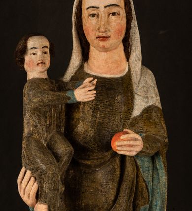 Zdjęcie nr 5: Stojąca figura Matki Boskiej z Dzieciątkiem reprezentuje przykład przyściennej rzeźby z tyłu wydrążonej. Maria stoi w lekkim kontrapoście, podkreślonym wysunięciem do przodu lewego kolana na niewielkim wzgórku. Prawą ręką podtrzymuje Dzieciątko, w lewej dłoni trzyma jabłko. Madonna ma mocno pociągłą twarz o jasnej karnacji z wysokim czołem, rumianych policzkach, długi, prosty nos i wydatne usta. Jej głowa osadzona jest na długiej szyi. Spod białej chusty ubranej na głowę i opadającej na ramiona widać brązowe włosy. Matka Boska na głowie ma koronę o ostrych, krótkich sterczynach. Nałożony na ciemnooliwkową suknię płaszcz w tym samym kolorze, podbity błękitem, tworzy dwa wałkowate zaokrąglone fałdy na brzuchu Marii, spływając miękkimi fałdami spod lewej ręki oraz festonem spod prawej ręki. Niżej fałdy płaszcza i sukni układają się niemal pionowo, opinając materią lewą nogę. Długa suknia odsłania czubki ciżemek. Dzieciątko zostało ujęte w trzech czwartych w prawo. Obie jego rączki wyciągnięte są przed siebie w stronę trzymanego przez Marię jabłka. Z kolei nóżki są skrzyżowane przy stopach. Jezus ma wyrazistą twarz o jasnej karnacji, małym nosem i wydatnych ustach. Jego głowę otaczają brązowe włosy układające się po bokach w charakterystyczne pukle. Jezus ubrany jest w długą, ciemnooliwkową tunikę odsłaniającą fragmenty stóp z charakterystycznymi mankietami przy rękawach. Szaty Matki Boskiej i Dzieciątka są srebrzone i laserowane.