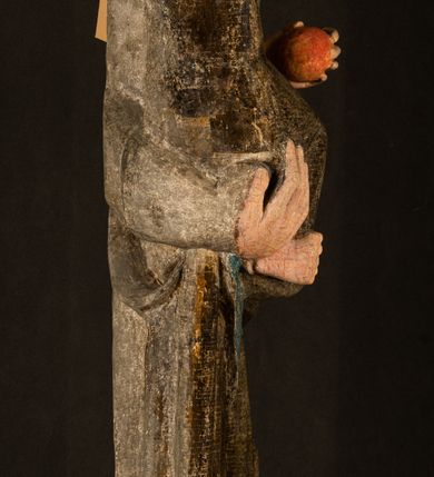 Zdjęcie nr 24: Stojąca figura Matki Boskiej z Dzieciątkiem reprezentuje przykład przyściennej rzeźby z tyłu wydrążonej. Maria stoi w lekkim kontrapoście, podkreślonym wysunięciem do przodu lewego kolana na niewielkim wzgórku. Prawą ręką podtrzymuje Dzieciątko, w lewej dłoni trzyma jabłko. Madonna ma mocno pociągłą twarz o jasnej karnacji z wysokim czołem, rumianych policzkach, długi, prosty nos i wydatne usta. Jej głowa osadzona jest na długiej szyi. Spod białej chusty ubranej na głowę i opadającej na ramiona widać brązowe włosy. Matka Boska na głowie ma koronę o ostrych, krótkich sterczynach. Nałożony na ciemnooliwkową suknię płaszcz w tym samym kolorze, podbity błękitem, tworzy dwa wałkowate zaokrąglone fałdy na brzuchu Marii, spływając miękkimi fałdami spod lewej ręki oraz festonem spod prawej ręki. Niżej fałdy płaszcza i sukni układają się niemal pionowo, opinając materią lewą nogę. Długa suknia odsłania czubki ciżemek. Dzieciątko zostało ujęte w trzech czwartych w prawo. Obie jego rączki wyciągnięte są przed siebie w stronę trzymanego przez Marię jabłka. Z kolei nóżki są skrzyżowane przy stopach. Jezus ma wyrazistą twarz o jasnej karnacji, małym nosem i wydatnych ustach. Jego głowę otaczają brązowe włosy układające się po bokach w charakterystyczne pukle. Jezus ubrany jest w długą, ciemnooliwkową tunikę odsłaniającą fragmenty stóp z charakterystycznymi mankietami przy rękawach. Szaty Matki Boskiej i Dzieciątka są srebrzone i laserowane.