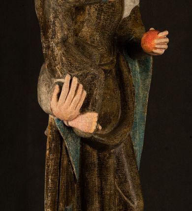 Zdjęcie nr 23: Stojąca figura Matki Boskiej z Dzieciątkiem reprezentuje przykład przyściennej rzeźby z tyłu wydrążonej. Maria stoi w lekkim kontrapoście, podkreślonym wysunięciem do przodu lewego kolana na niewielkim wzgórku. Prawą ręką podtrzymuje Dzieciątko, w lewej dłoni trzyma jabłko. Madonna ma mocno pociągłą twarz o jasnej karnacji z wysokim czołem, rumianych policzkach, długi, prosty nos i wydatne usta. Jej głowa osadzona jest na długiej szyi. Spod białej chusty ubranej na głowę i opadającej na ramiona widać brązowe włosy. Matka Boska na głowie ma koronę o ostrych, krótkich sterczynach. Nałożony na ciemnooliwkową suknię płaszcz w tym samym kolorze, podbity błękitem, tworzy dwa wałkowate zaokrąglone fałdy na brzuchu Marii, spływając miękkimi fałdami spod lewej ręki oraz festonem spod prawej ręki. Niżej fałdy płaszcza i sukni układają się niemal pionowo, opinając materią lewą nogę. Długa suknia odsłania czubki ciżemek. Dzieciątko zostało ujęte w trzech czwartych w prawo. Obie jego rączki wyciągnięte są przed siebie w stronę trzymanego przez Marię jabłka. Z kolei nóżki są skrzyżowane przy stopach. Jezus ma wyrazistą twarz o jasnej karnacji, małym nosem i wydatnych ustach. Jego głowę otaczają brązowe włosy układające się po bokach w charakterystyczne pukle. Jezus ubrany jest w długą, ciemnooliwkową tunikę odsłaniającą fragmenty stóp z charakterystycznymi mankietami przy rękawach. Szaty Matki Boskiej i Dzieciątka są srebrzone i laserowane.