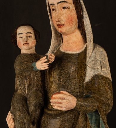 Zdjęcie nr 7: Stojąca figura Matki Boskiej z Dzieciątkiem reprezentuje przykład przyściennej rzeźby z tyłu wydrążonej. Maria stoi w lekkim kontrapoście, podkreślonym wysunięciem do przodu lewego kolana na niewielkim wzgórku. Prawą ręką podtrzymuje Dzieciątko, w lewej dłoni trzyma jabłko. Madonna ma mocno pociągłą twarz o jasnej karnacji z wysokim czołem, rumianych policzkach, długi, prosty nos i wydatne usta. Jej głowa osadzona jest na długiej szyi. Spod białej chusty ubranej na głowę i opadającej na ramiona widać brązowe włosy. Matka Boska na głowie ma koronę o ostrych, krótkich sterczynach. Nałożony na ciemnooliwkową suknię płaszcz w tym samym kolorze, podbity błękitem, tworzy dwa wałkowate zaokrąglone fałdy na brzuchu Marii, spływając miękkimi fałdami spod lewej ręki oraz festonem spod prawej ręki. Niżej fałdy płaszcza i sukni układają się niemal pionowo, opinając materią lewą nogę. Długa suknia odsłania czubki ciżemek. Dzieciątko zostało ujęte w trzech czwartych w prawo. Obie jego rączki wyciągnięte są przed siebie w stronę trzymanego przez Marię jabłka. Z kolei nóżki są skrzyżowane przy stopach. Jezus ma wyrazistą twarz o jasnej karnacji, małym nosem i wydatnych ustach. Jego głowę otaczają brązowe włosy układające się po bokach w charakterystyczne pukle. Jezus ubrany jest w długą, ciemnooliwkową tunikę odsłaniającą fragmenty stóp z charakterystycznymi mankietami przy rękawach. Szaty Matki Boskiej i Dzieciątka są srebrzone i laserowane.