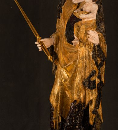 Zdjęcie nr 22: Rzeźba drążona ustawiona na niewielkim, wielobocznym postumencie. Maria ukazana w pozycji stojącej, frontalnie z ciałem ułożonym z lekkim kontrapoście. Lewą ręką podtrzymuje Dzieciątko Jezus, w prawej trzyma złocone berło zwieńczone kwiatem. Matka Boska przedstawiona jako młoda kobieta o podłużnej twarzy z wysokim czołem, migdałowatymi oczami oraz krótkim nosem i małymi ustami ułożonymi w delikatny uśmiech. Karnacja jasna, policzki wyraźnie zaróżowione. Długie, ciemne i falowane włosy rozdzielone przedziałkiem opadają na ramiona postaci. Na głowie Marii ciemna chusta oraz złocona korona (pozostałość korony?) lub diadem. Madonna jest ubrana w ciemną suknię z długimi rękawami dekorowaną motywem roślinnym oraz złocony płaszcz spięty pod szyją klamrą w kształcie kwiatu. Dzieciątko Jezus przedstawione w pozycji siedzącej z głową mocno pochyloną w bok i skrzyżowanymi stópkami. W ręku trzyma otwartą księgę. Twarz owalna, pulchna, ciemne oczy skierowane w lewo, usta w wyraźnym uśmiechu. Włosy, krótkie, falowane w kolorze brązowym. Dzieciątko jest ubrane w złoconą tkaninę udrapowaną wokół bioder. Figura polichromowana i częściowo złocona. Na odwrociu napis „Powtóre odzłocona / zarządem x(iędza) / Pawła Pomian Izdeb / skiego przez / Karol(a) Rośka / R(oku) P(ańskiego) 1853”.