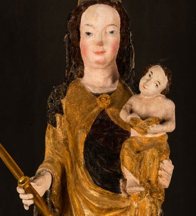 Zdjęcie nr 9: Rzeźba drążona ustawiona na niewielkim, wielobocznym postumencie. Maria ukazana w pozycji stojącej, frontalnie z ciałem ułożonym z lekkim kontrapoście. Lewą ręką podtrzymuje Dzieciątko Jezus, w prawej trzyma złocone berło zwieńczone kwiatem. Matka Boska przedstawiona jako młoda kobieta o podłużnej twarzy z wysokim czołem, migdałowatymi oczami oraz krótkim nosem i małymi ustami ułożonymi w delikatny uśmiech. Karnacja jasna, policzki wyraźnie zaróżowione. Długie, ciemne i falowane włosy rozdzielone przedziałkiem opadają na ramiona postaci. Na głowie Marii ciemna chusta oraz złocona korona (pozostałość korony?) lub diadem. Madonna jest ubrana w ciemną suknię z długimi rękawami dekorowaną motywem roślinnym oraz złocony płaszcz spięty pod szyją klamrą w kształcie kwiatu. Dzieciątko Jezus przedstawione w pozycji siedzącej z głową mocno pochyloną w bok i skrzyżowanymi stópkami. W ręku trzyma otwartą księgę. Twarz owalna, pulchna, ciemne oczy skierowane w lewo, usta w wyraźnym uśmiechu. Włosy, krótkie, falowane w kolorze brązowym. Dzieciątko jest ubrane w złoconą tkaninę udrapowaną wokół bioder. Figura polichromowana i częściowo złocona. Na odwrociu napis „Powtóre odzłocona / zarządem x(iędza) / Pawła Pomian Izdeb / skiego przez / Karol(a) Rośka / R(oku) P(ańskiego) 1853”.