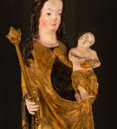 Zdjęcie nr 8: Rzeźba drążona ustawiona na niewielkim, wielobocznym postumencie. Maria ukazana w pozycji stojącej, frontalnie z ciałem ułożonym z lekkim kontrapoście. Lewą ręką podtrzymuje Dzieciątko Jezus, w prawej trzyma złocone berło zwieńczone kwiatem. Matka Boska przedstawiona jako młoda kobieta o podłużnej twarzy z wysokim czołem, migdałowatymi oczami oraz krótkim nosem i małymi ustami ułożonymi w delikatny uśmiech. Karnacja jasna, policzki wyraźnie zaróżowione. Długie, ciemne i falowane włosy rozdzielone przedziałkiem opadają na ramiona postaci. Na głowie Marii ciemna chusta oraz złocona korona (pozostałość korony?) lub diadem. Madonna jest ubrana w ciemną suknię z długimi rękawami dekorowaną motywem roślinnym oraz złocony płaszcz spięty pod szyją klamrą w kształcie kwiatu. Dzieciątko Jezus przedstawione w pozycji siedzącej z głową mocno pochyloną w bok i skrzyżowanymi stópkami. W ręku trzyma otwartą księgę. Twarz owalna, pulchna, ciemne oczy skierowane w lewo, usta w wyraźnym uśmiechu. Włosy, krótkie, falowane w kolorze brązowym. Dzieciątko jest ubrane w złoconą tkaninę udrapowaną wokół bioder. Figura polichromowana i częściowo złocona. Na odwrociu napis „Powtóre odzłocona / zarządem x(iędza) / Pawła Pomian Izdeb / skiego przez / Karol(a) Rośka / R(oku) P(ańskiego) 1853”.