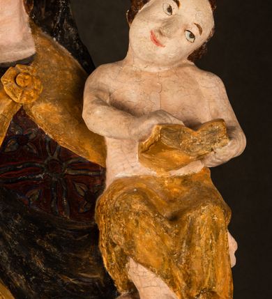 Zdjęcie nr 18: Rzeźba drążona ustawiona na niewielkim, wielobocznym postumencie. Maria ukazana w pozycji stojącej, frontalnie z ciałem ułożonym z lekkim kontrapoście. Lewą ręką podtrzymuje Dzieciątko Jezus, w prawej trzyma złocone berło zwieńczone kwiatem. Matka Boska przedstawiona jako młoda kobieta o podłużnej twarzy z wysokim czołem, migdałowatymi oczami oraz krótkim nosem i małymi ustami ułożonymi w delikatny uśmiech. Karnacja jasna, policzki wyraźnie zaróżowione. Długie, ciemne i falowane włosy rozdzielone przedziałkiem opadają na ramiona postaci. Na głowie Marii ciemna chusta oraz złocona korona (pozostałość korony?) lub diadem. Madonna jest ubrana w ciemną suknię z długimi rękawami dekorowaną motywem roślinnym oraz złocony płaszcz spięty pod szyją klamrą w kształcie kwiatu. Dzieciątko Jezus przedstawione w pozycji siedzącej z głową mocno pochyloną w bok i skrzyżowanymi stópkami. W ręku trzyma otwartą księgę. Twarz owalna, pulchna, ciemne oczy skierowane w lewo, usta w wyraźnym uśmiechu. Włosy, krótkie, falowane w kolorze brązowym. Dzieciątko jest ubrane w złoconą tkaninę udrapowaną wokół bioder. Figura polichromowana i częściowo złocona. Na odwrociu napis „Powtóre odzłocona / zarządem x(iędza) / Pawła Pomian Izdeb / skiego przez / Karol(a) Rośka / R(oku) P(ańskiego) 1853”.