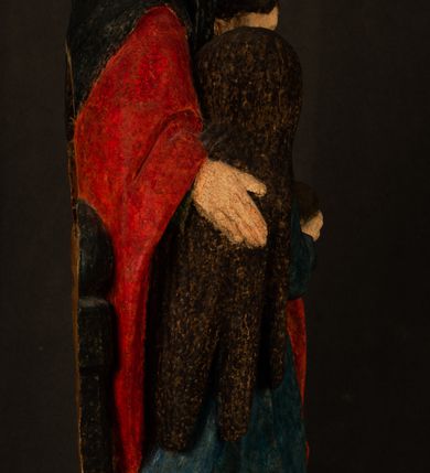 Zdjęcie nr 12: Rzeźba drążona przedstawiająca siedzącą św. Annę w towarzystwie klęczącej Marii oraz Dzieciątka Jezus. Anna ukazana frontalnie, z głową pochyloną nieznacznie w dół, obejmująca rękoma przedstawioną z prawej strony Marię oraz znajdujące się po lewej stronie Dzieciątko Jezus. Twarz świętej owalna o spokojnym wyrazie, z oczami skierowanymi w dół. Nos długi, prosty, na policzkach zaznaczony delikatny rumieniec. Anna ubrana jest w ciemnozieloną suknię przepasaną w talii, ciemnogranatową chustę zarzuconą na głowę, pod którą znajduje się biała podwika. Na ramiona i kolana postaci narzucony jest obszerny, czerwony płaszcz spod, którego widoczny jest ostro zakończony czubek brązowego buta św. Anny. Maria znajdująca się z lewej strony kompozycji została  przedstawiona jako młoda kobieta, klęcząca u stóp swojej matki i zwracająca się w kierunku siedzącego na kolanach Anny Dzieciątka Jezus. Owalną twarz Marii charakteryzuje wysokie czoło i cienkie, łukowato wygięte brwi. Długie, ciemne i lekko falowane włosy opadają na ramiona i plecy postaci. Maria swoją prawą rękę wspiera na kolanach Anny, lewą wyciąga przed siebie, podając Dzieciątku jabłko królewskie. Ubrana jest w ciemnogranatową suknię z długimi rękawami, spod której widoczne są bose stopy. Chrystus został ukazany jako nagi, ciemnowłosy chłopiec  o okrągłej twarzy z rumieńcem siedzący na kolanach św. Anny i wyciągający obie ręce w kierunku Marii. Rzeźba w pełni polichromowana.