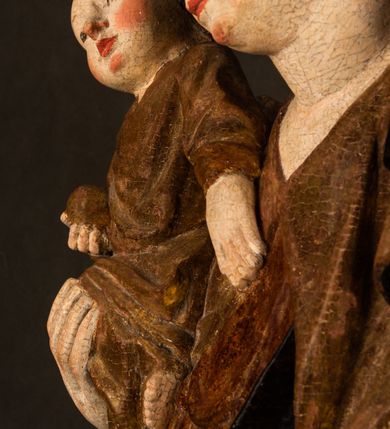 Zdjęcie nr 11: Przyścienna rzeźba Madonny z Dzieciątkiem jest z tyłu wydrążona. W tyle głowy Marii wbity jest hak służący do umocowania w ołtarzu. Matka Boska w całej postaci została ukazana w lekkim kontrapoście z charakterystycznym przegięciem w kształcie odwróconej litery "S". Na prawej ręce trzyma Dzieciątko Jezus, lewą zaś ręką opuszczoną wzdłuż tułowia podtrzymuje nadgarstkiem fałdy płaszcza. Madonna ma pociągłą, zamyśloną twarz o jasnej karnacji, wysokie czoło i pucułowate, rumiane policzki. Ma prosty nos, migdałowe oczy oraz wydatne usta i podbródek. Jej głowę osadzoną na długiej szyi okalają zaczesane do tyłu brązowe włosy, widoczne spod osuwającej się chusty opadającej na ramiona dwoma festonami. Maria ubrana jest w złoconą suknię laserowaną ciepłą czerwienią oraz złocony płaszcz podbity srebrną podszewką z oliwkowo zielonym laserunkiem. Z przodu płaszcz drapowany jest w łukowate fałdy. Jedna z nich biegnie diagonalnie od prawej dłoni ku lewej stopie Madonny. Płaszcz po bokach postaci spływa rurkowatymi kaskadami. Maria na głowię ma wysoką, złotą koronę ze sterczynami o trójlistnych zakończeniach. Mały Jezus został ujęty w trzech czwartych z nieznacznie odchyloną głową do tyłu i skrzyżowanymi nóżkami. Lewą rękę kładzie na piersi matki, w prawej zaś trzyma złocone jabłko. Ma okrągłą, pucułowatą twarz o rumianych policzkach. Charakteryzuje ją prosty nosek, wydatne wargi i podbródek oraz wyraziste, szeroko otwarte oczy. Jego głowę z wysokim czołem okalają krótkie brązowe włosy. Ubrany jest w złocistą długą tunikę odsłaniającą fragmenty stóp.   