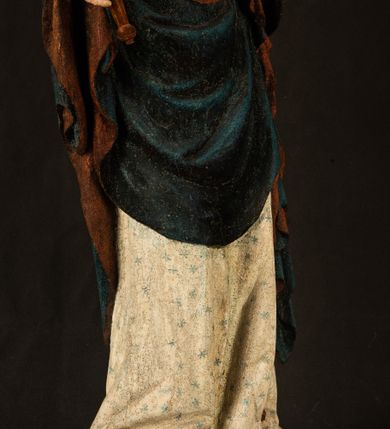 Zdjęcie nr 24: Rzeźba drążona, ukazująca Matkę Boską z Dzieciątkiem. Maria ustawiona frontalnie, w delikatnym kontrapoście, na lewym ręku trzyma Dzieciątko, w prawym dzierży berło. Twarz okrągła z wysokim czołem, drobnymi ustami i małymi oczami o delikatnie zarysowanych łukach brwiowych. Włosy jasne, ułożone w skręcone pukle spływające na plecy. Maria ubrana jest w białą suknię zdobioną drobnymi, niebieskimi gwiazdkami, ciemnobłękitny płaszcz z bordową podszewką spięty na piersi oraz spiczaste, brązowe buty. Na głowie ma założony ciemnoniebieski welon z bordową podszewką, spływający na ramiona i piersi oraz brązową podstawę do niezachowanej korony. Draperia szat miękka o wyraźnie zaznaczonych, licznych fałdach misowych i kaskadowych. Dzieciątko ukazane w pozycji siedzącej, zwrócone w prawo. W lewej dłoni trzyma jabłko królewskie, a prawą wyprostowaną podnosi na wysokość tułowia. Twarz pełna o dziecięcych rysach z małymi ustami, okolona krótkimi i kędzierzawymi włosami. Polichromia w odsłoniętych partiach ciała naturalistyczna, berło polichromowane na brązowo.