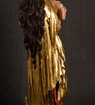 Zdjęcie nr 18: Całopostaciowa, pełnoplastyczna figura Madonny z Dzieciątkiem stojącej w wydatnym kontrapoście na podstawie o wykroju w kształcie półkola z parą wklęsek. Sylwetkę Marii charakteryzuje S-kształtny układ. Zwróconą w trzech czwartych w prawo, lekko przechyloną w bok głowę charakteryzują długie, ciemne, kręcone włosy opadające na plecy i lewe ramię oraz pociągła, owalna twarz o bardzo wysokim czole, łagodnych lukach ciemnych brwi, migdałowatych oczach, długim, zaokrąglonym na końcu nosie, pełnych, zaróżowionych policzkach, kształtnych ustach i zaokrąglonym podbródku. Długą szyję uwydatnia głęboki, półkolisty dekolt długiej, czerwonej sukni ze srebrnymi lamówkami, o długich, wąskich rękawach i ściśle przylegającej do tułowia. Sylwetkę ciasno osłania złocony płaszcz narzucony na prawe ramię osłaniając rękę talię i nogi do wysokości łydek. Spod sukni widoczne są ciemnobrązowe, spiczaste buty. Draperie cechuje dekoracyjny układ fałdowań w postaci spiętrzonej kaskady drobnych fałd rurkowych wzdłuż prawego biodra i nogi Marii oraz 3 promieniście opadających, C-kształtnych załamań. Nieco masywniejsze fałdy w dolnej partii sukni załamują się ostrymi formami pod wpływem styku z obuwiem i podłożem. Pod stopami sierp księżyca.  Maria prawą ręką podtrzymuje siedzące, zwrócone w trzech czwartych w lewo Dzieciątko lewą dłonią ujmując jego stopy. Jego nagą sylwetkę charakteryzuje stosunkowo sumaryczny modelunek ciała. Lekko odchyloną do tyłu głowę charakteryzuje owalna twarz o pełnych, zaokrąglonych rysach, z lekko wypukłym czołem, wyrazistymi łukami ciemnych, opadających brwi, dużymi, ciemnymi oczami, krótkim, lekko zadartym nosem, pełnymi ustami, zaróżowionymi policzkami i wyraźnie zarysowanym podbródkiem. Włosy krótkie, ciemne, mocno skręcone z wyraźnym puklem nad czołem, szyja krótka i szeroka. Zgięte w łokciach ręce uniesione są na wysokości klatki piersiowej, prawa bliżej sylwetki w geście błogosławieństwa, lewa nieco wysunięta do przodu z dłonią odwróconą ku górze. Prawa noga wyprostowana, lewa lekko zgięta w kolanie.       