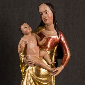 Zdjęcie nr 1: Całopostaciowa, pełnoplastyczna figura Madonny z Dzieciątkiem stojącej w wydatnym kontrapoście na podstawie o wykroju w kształcie półkola z parą wklęsek. Sylwetkę Marii charakteryzuje S-kształtny układ. Zwróconą w trzech czwartych w prawo, lekko przechyloną w bok głowę charakteryzują długie, ciemne, kręcone włosy opadające na plecy i lewe ramię oraz pociągła, owalna twarz o bardzo wysokim czole, łagodnych lukach ciemnych brwi, migdałowatych oczach, długim, zaokrąglonym na końcu nosie, pełnych, zaróżowionych policzkach, kształtnych ustach i zaokrąglonym podbródku. Długą szyję uwydatnia głęboki, półkolisty dekolt długiej, czerwonej sukni ze srebrnymi lamówkami, o długich, wąskich rękawach i ściśle przylegającej do tułowia. Sylwetkę ciasno osłania złocony płaszcz narzucony na prawe ramię osłaniając rękę talię i nogi do wysokości łydek. Spod sukni widoczne są ciemnobrązowe, spiczaste buty. Draperie cechuje dekoracyjny układ fałdowań w postaci spiętrzonej kaskady drobnych fałd rurkowych wzdłuż prawego biodra i nogi Marii oraz 3 promieniście opadających, C-kształtnych załamań. Nieco masywniejsze fałdy w dolnej partii sukni załamują się ostrymi formami pod wpływem styku z obuwiem i podłożem. Pod stopami sierp księżyca.  Maria prawą ręką podtrzymuje siedzące, zwrócone w trzech czwartych w lewo Dzieciątko lewą dłonią ujmując jego stopy. Jego nagą sylwetkę charakteryzuje stosunkowo sumaryczny modelunek ciała. Lekko odchyloną do tyłu głowę charakteryzuje owalna twarz o pełnych, zaokrąglonych rysach, z lekko wypukłym czołem, wyrazistymi łukami ciemnych, opadających brwi, dużymi, ciemnymi oczami, krótkim, lekko zadartym nosem, pełnymi ustami, zaróżowionymi policzkami i wyraźnie zarysowanym podbródkiem. Włosy krótkie, ciemne, mocno skręcone z wyraźnym puklem nad czołem, szyja krótka i szeroka. Zgięte w łokciach ręce uniesione są na wysokości klatki piersiowej, prawa bliżej sylwetki w geście błogosławieństwa, lewa nieco wysunięta do przodu z dłonią odwróconą ku górze. Prawa noga wyprostowana, lewa lekko zgięta w kolanie.       