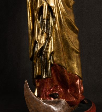 Zdjęcie nr 20: Całopostaciowa, pełnoplastyczna figura Madonny z Dzieciątkiem stojącej w wydatnym kontrapoście na podstawie o wykroju w kształcie półkola z parą wklęsek. Sylwetkę Marii charakteryzuje S-kształtny układ. Zwróconą w trzech czwartych w prawo, lekko przechyloną w bok głowę charakteryzują długie, ciemne, kręcone włosy opadające na plecy i lewe ramię oraz pociągła, owalna twarz o bardzo wysokim czole, łagodnych lukach ciemnych brwi, migdałowatych oczach, długim, zaokrąglonym na końcu nosie, pełnych, zaróżowionych policzkach, kształtnych ustach i zaokrąglonym podbródku. Długą szyję uwydatnia głęboki, półkolisty dekolt długiej, czerwonej sukni ze srebrnymi lamówkami, o długich, wąskich rękawach i ściśle przylegającej do tułowia. Sylwetkę ciasno osłania złocony płaszcz narzucony na prawe ramię osłaniając rękę talię i nogi do wysokości łydek. Spod sukni widoczne są ciemnobrązowe, spiczaste buty. Draperie cechuje dekoracyjny układ fałdowań w postaci spiętrzonej kaskady drobnych fałd rurkowych wzdłuż prawego biodra i nogi Marii oraz 3 promieniście opadających, C-kształtnych załamań. Nieco masywniejsze fałdy w dolnej partii sukni załamują się ostrymi formami pod wpływem styku z obuwiem i podłożem. Pod stopami sierp księżyca.  Maria prawą ręką podtrzymuje siedzące, zwrócone w trzech czwartych w lewo Dzieciątko lewą dłonią ujmując jego stopy. Jego nagą sylwetkę charakteryzuje stosunkowo sumaryczny modelunek ciała. Lekko odchyloną do tyłu głowę charakteryzuje owalna twarz o pełnych, zaokrąglonych rysach, z lekko wypukłym czołem, wyrazistymi łukami ciemnych, opadających brwi, dużymi, ciemnymi oczami, krótkim, lekko zadartym nosem, pełnymi ustami, zaróżowionymi policzkami i wyraźnie zarysowanym podbródkiem. Włosy krótkie, ciemne, mocno skręcone z wyraźnym puklem nad czołem, szyja krótka i szeroka. Zgięte w łokciach ręce uniesione są na wysokości klatki piersiowej, prawa bliżej sylwetki w geście błogosławieństwa, lewa nieco wysunięta do przodu z dłonią odwróconą ku górze. Prawa noga wyprostowana, lewa lekko zgięta w kolanie.       