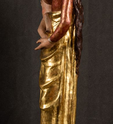 Zdjęcie nr 15: Całopostaciowa, pełnoplastyczna figura Madonny z Dzieciątkiem stojącej w wydatnym kontrapoście na podstawie o wykroju w kształcie półkola z parą wklęsek. Sylwetkę Marii charakteryzuje S-kształtny układ. Zwróconą w trzech czwartych w prawo, lekko przechyloną w bok głowę charakteryzują długie, ciemne, kręcone włosy opadające na plecy i lewe ramię oraz pociągła, owalna twarz o bardzo wysokim czole, łagodnych lukach ciemnych brwi, migdałowatych oczach, długim, zaokrąglonym na końcu nosie, pełnych, zaróżowionych policzkach, kształtnych ustach i zaokrąglonym podbródku. Długą szyję uwydatnia głęboki, półkolisty dekolt długiej, czerwonej sukni ze srebrnymi lamówkami, o długich, wąskich rękawach i ściśle przylegającej do tułowia. Sylwetkę ciasno osłania złocony płaszcz narzucony na prawe ramię osłaniając rękę talię i nogi do wysokości łydek. Spod sukni widoczne są ciemnobrązowe, spiczaste buty. Draperie cechuje dekoracyjny układ fałdowań w postaci spiętrzonej kaskady drobnych fałd rurkowych wzdłuż prawego biodra i nogi Marii oraz 3 promieniście opadających, C-kształtnych załamań. Nieco masywniejsze fałdy w dolnej partii sukni załamują się ostrymi formami pod wpływem styku z obuwiem i podłożem. Pod stopami sierp księżyca.  Maria prawą ręką podtrzymuje siedzące, zwrócone w trzech czwartych w lewo Dzieciątko lewą dłonią ujmując jego stopy. Jego nagą sylwetkę charakteryzuje stosunkowo sumaryczny modelunek ciała. Lekko odchyloną do tyłu głowę charakteryzuje owalna twarz o pełnych, zaokrąglonych rysach, z lekko wypukłym czołem, wyrazistymi łukami ciemnych, opadających brwi, dużymi, ciemnymi oczami, krótkim, lekko zadartym nosem, pełnymi ustami, zaróżowionymi policzkami i wyraźnie zarysowanym podbródkiem. Włosy krótkie, ciemne, mocno skręcone z wyraźnym puklem nad czołem, szyja krótka i szeroka. Zgięte w łokciach ręce uniesione są na wysokości klatki piersiowej, prawa bliżej sylwetki w geście błogosławieństwa, lewa nieco wysunięta do przodu z dłonią odwróconą ku górze. Prawa noga wyprostowana, lewa lekko zgięta w kolanie.       