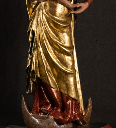 Zdjęcie nr 19: Całopostaciowa, pełnoplastyczna figura Madonny z Dzieciątkiem stojącej w wydatnym kontrapoście na podstawie o wykroju w kształcie półkola z parą wklęsek. Sylwetkę Marii charakteryzuje S-kształtny układ. Zwróconą w trzech czwartych w prawo, lekko przechyloną w bok głowę charakteryzują długie, ciemne, kręcone włosy opadające na plecy i lewe ramię oraz pociągła, owalna twarz o bardzo wysokim czole, łagodnych lukach ciemnych brwi, migdałowatych oczach, długim, zaokrąglonym na końcu nosie, pełnych, zaróżowionych policzkach, kształtnych ustach i zaokrąglonym podbródku. Długą szyję uwydatnia głęboki, półkolisty dekolt długiej, czerwonej sukni ze srebrnymi lamówkami, o długich, wąskich rękawach i ściśle przylegającej do tułowia. Sylwetkę ciasno osłania złocony płaszcz narzucony na prawe ramię osłaniając rękę talię i nogi do wysokości łydek. Spod sukni widoczne są ciemnobrązowe, spiczaste buty. Draperie cechuje dekoracyjny układ fałdowań w postaci spiętrzonej kaskady drobnych fałd rurkowych wzdłuż prawego biodra i nogi Marii oraz 3 promieniście opadających, C-kształtnych załamań. Nieco masywniejsze fałdy w dolnej partii sukni załamują się ostrymi formami pod wpływem styku z obuwiem i podłożem. Pod stopami sierp księżyca.  Maria prawą ręką podtrzymuje siedzące, zwrócone w trzech czwartych w lewo Dzieciątko lewą dłonią ujmując jego stopy. Jego nagą sylwetkę charakteryzuje stosunkowo sumaryczny modelunek ciała. Lekko odchyloną do tyłu głowę charakteryzuje owalna twarz o pełnych, zaokrąglonych rysach, z lekko wypukłym czołem, wyrazistymi łukami ciemnych, opadających brwi, dużymi, ciemnymi oczami, krótkim, lekko zadartym nosem, pełnymi ustami, zaróżowionymi policzkami i wyraźnie zarysowanym podbródkiem. Włosy krótkie, ciemne, mocno skręcone z wyraźnym puklem nad czołem, szyja krótka i szeroka. Zgięte w łokciach ręce uniesione są na wysokości klatki piersiowej, prawa bliżej sylwetki w geście błogosławieństwa, lewa nieco wysunięta do przodu z dłonią odwróconą ku górze. Prawa noga wyprostowana, lewa lekko zgięta w kolanie.       