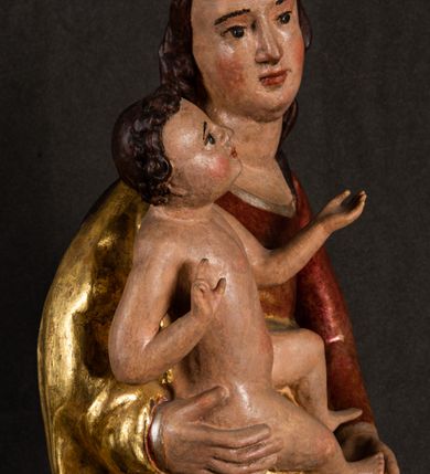 Zdjęcie nr 6: Całopostaciowa, pełnoplastyczna figura Madonny z Dzieciątkiem stojącej w wydatnym kontrapoście na podstawie o wykroju w kształcie półkola z parą wklęsek. Sylwetkę Marii charakteryzuje S-kształtny układ. Zwróconą w trzech czwartych w prawo, lekko przechyloną w bok głowę charakteryzują długie, ciemne, kręcone włosy opadające na plecy i lewe ramię oraz pociągła, owalna twarz o bardzo wysokim czole, łagodnych lukach ciemnych brwi, migdałowatych oczach, długim, zaokrąglonym na końcu nosie, pełnych, zaróżowionych policzkach, kształtnych ustach i zaokrąglonym podbródku. Długą szyję uwydatnia głęboki, półkolisty dekolt długiej, czerwonej sukni ze srebrnymi lamówkami, o długich, wąskich rękawach i ściśle przylegającej do tułowia. Sylwetkę ciasno osłania złocony płaszcz narzucony na prawe ramię osłaniając rękę talię i nogi do wysokości łydek. Spod sukni widoczne są ciemnobrązowe, spiczaste buty. Draperie cechuje dekoracyjny układ fałdowań w postaci spiętrzonej kaskady drobnych fałd rurkowych wzdłuż prawego biodra i nogi Marii oraz 3 promieniście opadających, C-kształtnych załamań. Nieco masywniejsze fałdy w dolnej partii sukni załamują się ostrymi formami pod wpływem styku z obuwiem i podłożem. Pod stopami sierp księżyca.  Maria prawą ręką podtrzymuje siedzące, zwrócone w trzech czwartych w lewo Dzieciątko lewą dłonią ujmując jego stopy. Jego nagą sylwetkę charakteryzuje stosunkowo sumaryczny modelunek ciała. Lekko odchyloną do tyłu głowę charakteryzuje owalna twarz o pełnych, zaokrąglonych rysach, z lekko wypukłym czołem, wyrazistymi łukami ciemnych, opadających brwi, dużymi, ciemnymi oczami, krótkim, lekko zadartym nosem, pełnymi ustami, zaróżowionymi policzkami i wyraźnie zarysowanym podbródkiem. Włosy krótkie, ciemne, mocno skręcone z wyraźnym puklem nad czołem, szyja krótka i szeroka. Zgięte w łokciach ręce uniesione są na wysokości klatki piersiowej, prawa bliżej sylwetki w geście błogosławieństwa, lewa nieco wysunięta do przodu z dłonią odwróconą ku górze. Prawa noga wyprostowana, lewa lekko zgięta w kolanie.       