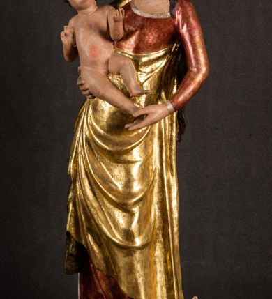 Zdjęcie nr 2: Całopostaciowa, pełnoplastyczna figura Madonny z Dzieciątkiem stojącej w wydatnym kontrapoście na podstawie o wykroju w kształcie półkola z parą wklęsek. Sylwetkę Marii charakteryzuje S-kształtny układ. Zwróconą w trzech czwartych w prawo, lekko przechyloną w bok głowę charakteryzują długie, ciemne, kręcone włosy opadające na plecy i lewe ramię oraz pociągła, owalna twarz o bardzo wysokim czole, łagodnych lukach ciemnych brwi, migdałowatych oczach, długim, zaokrąglonym na końcu nosie, pełnych, zaróżowionych policzkach, kształtnych ustach i zaokrąglonym podbródku. Długą szyję uwydatnia głęboki, półkolisty dekolt długiej, czerwonej sukni ze srebrnymi lamówkami, o długich, wąskich rękawach i ściśle przylegającej do tułowia. Sylwetkę ciasno osłania złocony płaszcz narzucony na prawe ramię osłaniając rękę talię i nogi do wysokości łydek. Spod sukni widoczne są ciemnobrązowe, spiczaste buty. Draperie cechuje dekoracyjny układ fałdowań w postaci spiętrzonej kaskady drobnych fałd rurkowych wzdłuż prawego biodra i nogi Marii oraz 3 promieniście opadających, C-kształtnych załamań. Nieco masywniejsze fałdy w dolnej partii sukni załamują się ostrymi formami pod wpływem styku z obuwiem i podłożem. Pod stopami sierp księżyca.  Maria prawą ręką podtrzymuje siedzące, zwrócone w trzech czwartych w lewo Dzieciątko lewą dłonią ujmując jego stopy. Jego nagą sylwetkę charakteryzuje stosunkowo sumaryczny modelunek ciała. Lekko odchyloną do tyłu głowę charakteryzuje owalna twarz o pełnych, zaokrąglonych rysach, z lekko wypukłym czołem, wyrazistymi łukami ciemnych, opadających brwi, dużymi, ciemnymi oczami, krótkim, lekko zadartym nosem, pełnymi ustami, zaróżowionymi policzkami i wyraźnie zarysowanym podbródkiem. Włosy krótkie, ciemne, mocno skręcone z wyraźnym puklem nad czołem, szyja krótka i szeroka. Zgięte w łokciach ręce uniesione są na wysokości klatki piersiowej, prawa bliżej sylwetki w geście błogosławieństwa, lewa nieco wysunięta do przodu z dłonią odwróconą ku górze. Prawa noga wyprostowana, lewa lekko zgięta w kolanie.       