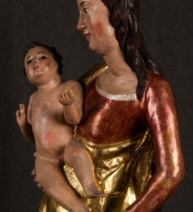 Zdjęcie nr 5: Całopostaciowa, pełnoplastyczna figura Madonny z Dzieciątkiem stojącej w wydatnym kontrapoście na podstawie o wykroju w kształcie półkola z parą wklęsek. Sylwetkę Marii charakteryzuje S-kształtny układ. Zwróconą w trzech czwartych w prawo, lekko przechyloną w bok głowę charakteryzują długie, ciemne, kręcone włosy opadające na plecy i lewe ramię oraz pociągła, owalna twarz o bardzo wysokim czole, łagodnych lukach ciemnych brwi, migdałowatych oczach, długim, zaokrąglonym na końcu nosie, pełnych, zaróżowionych policzkach, kształtnych ustach i zaokrąglonym podbródku. Długą szyję uwydatnia głęboki, półkolisty dekolt długiej, czerwonej sukni ze srebrnymi lamówkami, o długich, wąskich rękawach i ściśle przylegającej do tułowia. Sylwetkę ciasno osłania złocony płaszcz narzucony na prawe ramię osłaniając rękę talię i nogi do wysokości łydek. Spod sukni widoczne są ciemnobrązowe, spiczaste buty. Draperie cechuje dekoracyjny układ fałdowań w postaci spiętrzonej kaskady drobnych fałd rurkowych wzdłuż prawego biodra i nogi Marii oraz 3 promieniście opadających, C-kształtnych załamań. Nieco masywniejsze fałdy w dolnej partii sukni załamują się ostrymi formami pod wpływem styku z obuwiem i podłożem. Pod stopami sierp księżyca.  Maria prawą ręką podtrzymuje siedzące, zwrócone w trzech czwartych w lewo Dzieciątko lewą dłonią ujmując jego stopy. Jego nagą sylwetkę charakteryzuje stosunkowo sumaryczny modelunek ciała. Lekko odchyloną do tyłu głowę charakteryzuje owalna twarz o pełnych, zaokrąglonych rysach, z lekko wypukłym czołem, wyrazistymi łukami ciemnych, opadających brwi, dużymi, ciemnymi oczami, krótkim, lekko zadartym nosem, pełnymi ustami, zaróżowionymi policzkami i wyraźnie zarysowanym podbródkiem. Włosy krótkie, ciemne, mocno skręcone z wyraźnym puklem nad czołem, szyja krótka i szeroka. Zgięte w łokciach ręce uniesione są na wysokości klatki piersiowej, prawa bliżej sylwetki w geście błogosławieństwa, lewa nieco wysunięta do przodu z dłonią odwróconą ku górze. Prawa noga wyprostowana, lewa lekko zgięta w kolanie.       