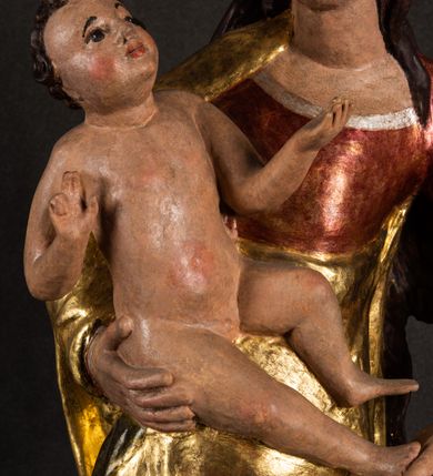 Zdjęcie nr 8: Całopostaciowa, pełnoplastyczna figura Madonny z Dzieciątkiem stojącej w wydatnym kontrapoście na podstawie o wykroju w kształcie półkola z parą wklęsek. Sylwetkę Marii charakteryzuje S-kształtny układ. Zwróconą w trzech czwartych w prawo, lekko przechyloną w bok głowę charakteryzują długie, ciemne, kręcone włosy opadające na plecy i lewe ramię oraz pociągła, owalna twarz o bardzo wysokim czole, łagodnych lukach ciemnych brwi, migdałowatych oczach, długim, zaokrąglonym na końcu nosie, pełnych, zaróżowionych policzkach, kształtnych ustach i zaokrąglonym podbródku. Długą szyję uwydatnia głęboki, półkolisty dekolt długiej, czerwonej sukni ze srebrnymi lamówkami, o długich, wąskich rękawach i ściśle przylegającej do tułowia. Sylwetkę ciasno osłania złocony płaszcz narzucony na prawe ramię osłaniając rękę talię i nogi do wysokości łydek. Spod sukni widoczne są ciemnobrązowe, spiczaste buty. Draperie cechuje dekoracyjny układ fałdowań w postaci spiętrzonej kaskady drobnych fałd rurkowych wzdłuż prawego biodra i nogi Marii oraz 3 promieniście opadających, C-kształtnych załamań. Nieco masywniejsze fałdy w dolnej partii sukni załamują się ostrymi formami pod wpływem styku z obuwiem i podłożem. Pod stopami sierp księżyca.  Maria prawą ręką podtrzymuje siedzące, zwrócone w trzech czwartych w lewo Dzieciątko lewą dłonią ujmując jego stopy. Jego nagą sylwetkę charakteryzuje stosunkowo sumaryczny modelunek ciała. Lekko odchyloną do tyłu głowę charakteryzuje owalna twarz o pełnych, zaokrąglonych rysach, z lekko wypukłym czołem, wyrazistymi łukami ciemnych, opadających brwi, dużymi, ciemnymi oczami, krótkim, lekko zadartym nosem, pełnymi ustami, zaróżowionymi policzkami i wyraźnie zarysowanym podbródkiem. Włosy krótkie, ciemne, mocno skręcone z wyraźnym puklem nad czołem, szyja krótka i szeroka. Zgięte w łokciach ręce uniesione są na wysokości klatki piersiowej, prawa bliżej sylwetki w geście błogosławieństwa, lewa nieco wysunięta do przodu z dłonią odwróconą ku górze. Prawa noga wyprostowana, lewa lekko zgięta w kolanie.       