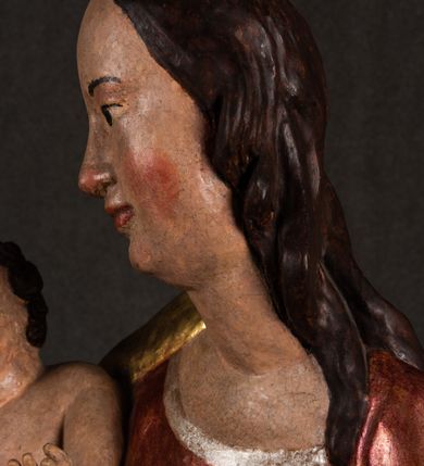 Zdjęcie nr 12: Całopostaciowa, pełnoplastyczna figura Madonny z Dzieciątkiem stojącej w wydatnym kontrapoście na podstawie o wykroju w kształcie półkola z parą wklęsek. Sylwetkę Marii charakteryzuje S-kształtny układ. Zwróconą w trzech czwartych w prawo, lekko przechyloną w bok głowę charakteryzują długie, ciemne, kręcone włosy opadające na plecy i lewe ramię oraz pociągła, owalna twarz o bardzo wysokim czole, łagodnych lukach ciemnych brwi, migdałowatych oczach, długim, zaokrąglonym na końcu nosie, pełnych, zaróżowionych policzkach, kształtnych ustach i zaokrąglonym podbródku. Długą szyję uwydatnia głęboki, półkolisty dekolt długiej, czerwonej sukni ze srebrnymi lamówkami, o długich, wąskich rękawach i ściśle przylegającej do tułowia. Sylwetkę ciasno osłania złocony płaszcz narzucony na prawe ramię osłaniając rękę talię i nogi do wysokości łydek. Spod sukni widoczne są ciemnobrązowe, spiczaste buty. Draperie cechuje dekoracyjny układ fałdowań w postaci spiętrzonej kaskady drobnych fałd rurkowych wzdłuż prawego biodra i nogi Marii oraz 3 promieniście opadających, C-kształtnych załamań. Nieco masywniejsze fałdy w dolnej partii sukni załamują się ostrymi formami pod wpływem styku z obuwiem i podłożem. Pod stopami sierp księżyca.  Maria prawą ręką podtrzymuje siedzące, zwrócone w trzech czwartych w lewo Dzieciątko lewą dłonią ujmując jego stopy. Jego nagą sylwetkę charakteryzuje stosunkowo sumaryczny modelunek ciała. Lekko odchyloną do tyłu głowę charakteryzuje owalna twarz o pełnych, zaokrąglonych rysach, z lekko wypukłym czołem, wyrazistymi łukami ciemnych, opadających brwi, dużymi, ciemnymi oczami, krótkim, lekko zadartym nosem, pełnymi ustami, zaróżowionymi policzkami i wyraźnie zarysowanym podbródkiem. Włosy krótkie, ciemne, mocno skręcone z wyraźnym puklem nad czołem, szyja krótka i szeroka. Zgięte w łokciach ręce uniesione są na wysokości klatki piersiowej, prawa bliżej sylwetki w geście błogosławieństwa, lewa nieco wysunięta do przodu z dłonią odwróconą ku górze. Prawa noga wyprostowana, lewa lekko zgięta w kolanie.       