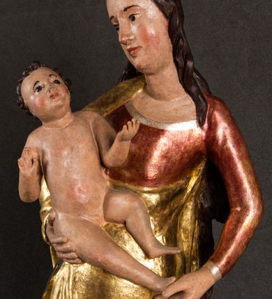 Zdjęcie nr 4: Całopostaciowa, pełnoplastyczna figura Madonny z Dzieciątkiem stojącej w wydatnym kontrapoście na podstawie o wykroju w kształcie półkola z parą wklęsek. Sylwetkę Marii charakteryzuje S-kształtny układ. Zwróconą w trzech czwartych w prawo, lekko przechyloną w bok głowę charakteryzują długie, ciemne, kręcone włosy opadające na plecy i lewe ramię oraz pociągła, owalna twarz o bardzo wysokim czole, łagodnych lukach ciemnych brwi, migdałowatych oczach, długim, zaokrąglonym na końcu nosie, pełnych, zaróżowionych policzkach, kształtnych ustach i zaokrąglonym podbródku. Długą szyję uwydatnia głęboki, półkolisty dekolt długiej, czerwonej sukni ze srebrnymi lamówkami, o długich, wąskich rękawach i ściśle przylegającej do tułowia. Sylwetkę ciasno osłania złocony płaszcz narzucony na prawe ramię osłaniając rękę talię i nogi do wysokości łydek. Spod sukni widoczne są ciemnobrązowe, spiczaste buty. Draperie cechuje dekoracyjny układ fałdowań w postaci spiętrzonej kaskady drobnych fałd rurkowych wzdłuż prawego biodra i nogi Marii oraz 3 promieniście opadających, C-kształtnych załamań. Nieco masywniejsze fałdy w dolnej partii sukni załamują się ostrymi formami pod wpływem styku z obuwiem i podłożem. Pod stopami sierp księżyca.  Maria prawą ręką podtrzymuje siedzące, zwrócone w trzech czwartych w lewo Dzieciątko lewą dłonią ujmując jego stopy. Jego nagą sylwetkę charakteryzuje stosunkowo sumaryczny modelunek ciała. Lekko odchyloną do tyłu głowę charakteryzuje owalna twarz o pełnych, zaokrąglonych rysach, z lekko wypukłym czołem, wyrazistymi łukami ciemnych, opadających brwi, dużymi, ciemnymi oczami, krótkim, lekko zadartym nosem, pełnymi ustami, zaróżowionymi policzkami i wyraźnie zarysowanym podbródkiem. Włosy krótkie, ciemne, mocno skręcone z wyraźnym puklem nad czołem, szyja krótka i szeroka. Zgięte w łokciach ręce uniesione są na wysokości klatki piersiowej, prawa bliżej sylwetki w geście błogosławieństwa, lewa nieco wysunięta do przodu z dłonią odwróconą ku górze. Prawa noga wyprostowana, lewa lekko zgięta w kolanie.       