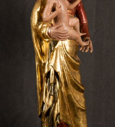 Zdjęcie nr 13: Całopostaciowa, pełnoplastyczna figura Madonny z Dzieciątkiem stojącej w wydatnym kontrapoście na podstawie o wykroju w kształcie półkola z parą wklęsek. Sylwetkę Marii charakteryzuje S-kształtny układ. Zwróconą w trzech czwartych w prawo, lekko przechyloną w bok głowę charakteryzują długie, ciemne, kręcone włosy opadające na plecy i lewe ramię oraz pociągła, owalna twarz o bardzo wysokim czole, łagodnych lukach ciemnych brwi, migdałowatych oczach, długim, zaokrąglonym na końcu nosie, pełnych, zaróżowionych policzkach, kształtnych ustach i zaokrąglonym podbródku. Długą szyję uwydatnia głęboki, półkolisty dekolt długiej, czerwonej sukni ze srebrnymi lamówkami, o długich, wąskich rękawach i ściśle przylegającej do tułowia. Sylwetkę ciasno osłania złocony płaszcz narzucony na prawe ramię osłaniając rękę talię i nogi do wysokości łydek. Spod sukni widoczne są ciemnobrązowe, spiczaste buty. Draperie cechuje dekoracyjny układ fałdowań w postaci spiętrzonej kaskady drobnych fałd rurkowych wzdłuż prawego biodra i nogi Marii oraz 3 promieniście opadających, C-kształtnych załamań. Nieco masywniejsze fałdy w dolnej partii sukni załamują się ostrymi formami pod wpływem styku z obuwiem i podłożem. Pod stopami sierp księżyca.  Maria prawą ręką podtrzymuje siedzące, zwrócone w trzech czwartych w lewo Dzieciątko lewą dłonią ujmując jego stopy. Jego nagą sylwetkę charakteryzuje stosunkowo sumaryczny modelunek ciała. Lekko odchyloną do tyłu głowę charakteryzuje owalna twarz o pełnych, zaokrąglonych rysach, z lekko wypukłym czołem, wyrazistymi łukami ciemnych, opadających brwi, dużymi, ciemnymi oczami, krótkim, lekko zadartym nosem, pełnymi ustami, zaróżowionymi policzkami i wyraźnie zarysowanym podbródkiem. Włosy krótkie, ciemne, mocno skręcone z wyraźnym puklem nad czołem, szyja krótka i szeroka. Zgięte w łokciach ręce uniesione są na wysokości klatki piersiowej, prawa bliżej sylwetki w geście błogosławieństwa, lewa nieco wysunięta do przodu z dłonią odwróconą ku górze. Prawa noga wyprostowana, lewa lekko zgięta w kolanie.       