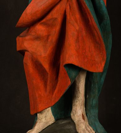 Zdjęcie nr 14: Pełnoplastyczna rzeźba ukazuje frontalnie w całej postaci zmartwychwstałego Chrystusa stojącego w kontrapoście na niewielkim wzgórku. Jego prawa noga jest nieco wysunięta i zgięta w kolanie. Zmartwychwstały unosi w geście zwycięstwa prawą rękę zgiętą w łokciu, lewą zaś uniesioną na wysokość piersi trzyma proporzec. Zbawiciel ma nieproporcjonalnie duże dłonie. Na rękach, stopach i boku widać ślady po męce. Chrystus ma mocno pociągłą twarz okoloną, krótkim, brązowym zarostem i brązowe włosy spływające na plecy. Jego twarz o jasnej karnacji charakteryzuje wysokie czoło, prosty nos i wydatne usta oraz szeroko otwarte ciemne oczy o spojrzeniu skierowanym przed siebie. Jezus ubrany jest w spięty pod szyją czerwony płaszcz podbity ciemną zielenią grynszpanową sięgający łydek z prawą połą podwiniętą przy lewym boku. Płaszcz odsłania mocno taliowany nagi tors z widocznym przebitym prawym bokiem.  