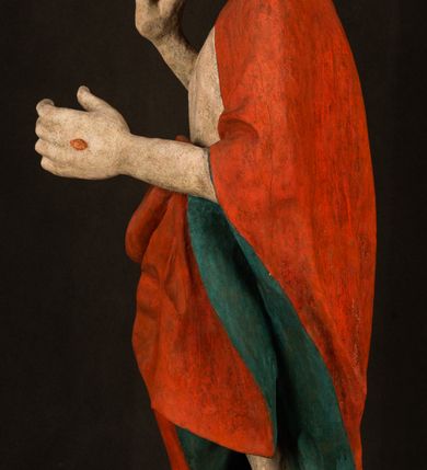 Zdjęcie nr 10: Pełnoplastyczna rzeźba ukazuje frontalnie w całej postaci zmartwychwstałego Chrystusa stojącego w kontrapoście na niewielkim wzgórku. Jego prawa noga jest nieco wysunięta i zgięta w kolanie. Zmartwychwstały unosi w geście zwycięstwa prawą rękę zgiętą w łokciu, lewą zaś uniesioną na wysokość piersi trzyma proporzec. Zbawiciel ma nieproporcjonalnie duże dłonie. Na rękach, stopach i boku widać ślady po męce. Chrystus ma mocno pociągłą twarz okoloną, krótkim, brązowym zarostem i brązowe włosy spływające na plecy. Jego twarz o jasnej karnacji charakteryzuje wysokie czoło, prosty nos i wydatne usta oraz szeroko otwarte ciemne oczy o spojrzeniu skierowanym przed siebie. Jezus ubrany jest w spięty pod szyją czerwony płaszcz podbity ciemną zielenią grynszpanową sięgający łydek z prawą połą podwiniętą przy lewym boku. Płaszcz odsłania mocno taliowany nagi tors z widocznym przebitym prawym bokiem.  