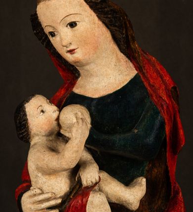 Zdjęcie nr 7: Rzeźba drążona, w pełni polichromowana. Pełnopostaciowe przedstawienie Marii karmiącej Dzieciątko Jezus. Matka Boska ukazana frontalnie, w pozycji siedzącej, trzyma na kolanach małego Jezusa, którego karmi piersią. Prawą ręką podtrzymuje Dzieciątko, lewą podaje mu pierś. Przedstawiona jako młoda kobieta o szerokiej, owalnej twarzy o wysokim czole i długich, ciemnych włosach. Oczy migdałowate, wpatrzone przed siebie. Cieńkie, łukowate brwi, niewielkie usta. Na głowie charakterystyczne podwyższenie – zapewne ślad po mocowaniu korony. Maria ubrana jest w ciemnogranatową, obcisłą suknię z długimi rękawami oraz czerwony płaszcz. Poła płaszcza okrywa również biodra Dzieciątka. Spod fałdów szat widoczne są czarne trzewiki o spiczastych noskach. Dzieciątko ukazane w pozycji siedzącej z głową uniesioną do góry, wyciągniętymi do przodu nóżkami i lewą ręką trzymającą pierś Marii. Twarz pełna z szeroko otwartymi oczami wpatrzonymi w matkę i zaróżowionymi policzkami. Włosy, krótkie, ciemne.