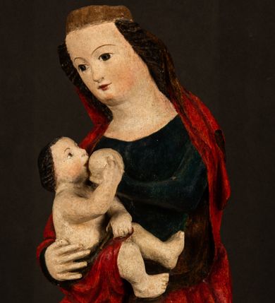 Zdjęcie nr 1: Rzeźba drążona, w pełni polichromowana. Pełnopostaciowe przedstawienie Marii karmiącej Dzieciątko Jezus. Matka Boska ukazana frontalnie, w pozycji siedzącej, trzyma na kolanach małego Jezusa, którego karmi piersią. Prawą ręką podtrzymuje Dzieciątko, lewą podaje mu pierś. Przedstawiona jako młoda kobieta o szerokiej, owalnej twarzy o wysokim czole i długich, ciemnych włosach. Oczy migdałowate, wpatrzone przed siebie. Cieńkie, łukowate brwi, niewielkie usta. Na głowie charakterystyczne podwyższenie – zapewne ślad po mocowaniu korony. Maria ubrana jest w ciemnogranatową, obcisłą suknię z długimi rękawami oraz czerwony płaszcz. Poła płaszcza okrywa również biodra Dzieciątka. Spod fałdów szat widoczne są czarne trzewiki o spiczastych noskach. Dzieciątko ukazane w pozycji siedzącej z głową uniesioną do góry, wyciągniętymi do przodu nóżkami i lewą ręką trzymającą pierś Marii. Twarz pełna z szeroko otwartymi oczami wpatrzonymi w matkę i zaróżowionymi policzkami. Włosy, krótkie, ciemne.