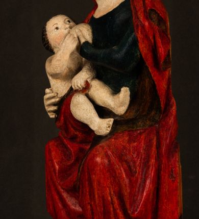 Zdjęcie nr 2: Rzeźba drążona, w pełni polichromowana. Pełnopostaciowe przedstawienie Marii karmiącej Dzieciątko Jezus. Matka Boska ukazana frontalnie, w pozycji siedzącej, trzyma na kolanach małego Jezusa, którego karmi piersią. Prawą ręką podtrzymuje Dzieciątko, lewą podaje mu pierś. Przedstawiona jako młoda kobieta o szerokiej, owalnej twarzy o wysokim czole i długich, ciemnych włosach. Oczy migdałowate, wpatrzone przed siebie. Cieńkie, łukowate brwi, niewielkie usta. Na głowie charakterystyczne podwyższenie – zapewne ślad po mocowaniu korony. Maria ubrana jest w ciemnogranatową, obcisłą suknię z długimi rękawami oraz czerwony płaszcz. Poła płaszcza okrywa również biodra Dzieciątka. Spod fałdów szat widoczne są czarne trzewiki o spiczastych noskach. Dzieciątko ukazane w pozycji siedzącej z głową uniesioną do góry, wyciągniętymi do przodu nóżkami i lewą ręką trzymającą pierś Marii. Twarz pełna z szeroko otwartymi oczami wpatrzonymi w matkę i zaróżowionymi policzkami. Włosy, krótkie, ciemne.