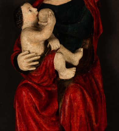 Zdjęcie nr 5: Rzeźba drążona, w pełni polichromowana. Pełnopostaciowe przedstawienie Marii karmiącej Dzieciątko Jezus. Matka Boska ukazana frontalnie, w pozycji siedzącej, trzyma na kolanach małego Jezusa, którego karmi piersią. Prawą ręką podtrzymuje Dzieciątko, lewą podaje mu pierś. Przedstawiona jako młoda kobieta o szerokiej, owalnej twarzy o wysokim czole i długich, ciemnych włosach. Oczy migdałowate, wpatrzone przed siebie. Cieńkie, łukowate brwi, niewielkie usta. Na głowie charakterystyczne podwyższenie – zapewne ślad po mocowaniu korony. Maria ubrana jest w ciemnogranatową, obcisłą suknię z długimi rękawami oraz czerwony płaszcz. Poła płaszcza okrywa również biodra Dzieciątka. Spod fałdów szat widoczne są czarne trzewiki o spiczastych noskach. Dzieciątko ukazane w pozycji siedzącej z głową uniesioną do góry, wyciągniętymi do przodu nóżkami i lewą ręką trzymającą pierś Marii. Twarz pełna z szeroko otwartymi oczami wpatrzonymi w matkę i zaróżowionymi policzkami. Włosy, krótkie, ciemne.