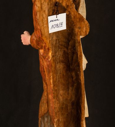 Zdjęcie nr 22: Rzeźba drążona, ustawiona na niewielkim, wielobocznym postumencie. Stojąca Maria ukazana frontalnie, w silnym kontrapoście z esowato wygiętym ciałem. Na lewej ręce podtrzymuje siedzące Dzieciątko, w prawej ręce ugiętej w łokciu i wyciągniętej do przodu najpewniej znajdowało się kiedyś berło.  Maria została ukazana jako młoda kobieta o owalnej twarzy, dużych, migdałowatych oczach, długim, prostym nosie i wyraźnie zaakcentowanych, dużych ustach. Karnacja jasna z rumieńcem zaznaczonym na policzkach i podbródku. Długie, jasne i falowane włosy częściowo zakryte chustą, symetrycznie okalają twarz postaci, spadając na jej ramiona. Maria jest ubrana w długą, sięgającą ziemi suknię, spod której widoczne są ostro zakończone czubki złotych butów oraz obszerny, złoty płaszcz tworzący ostro załamujące się fałdy na wysokości kolan Marii i formujący kaskadę wzdłuż prawego boku postaci. Na głowie Marii znajduje się biała chusta ze złota lamówką. Dzieciątko Jezus ukazane w pozycji siedzącej z lewą nogą wysuniętą wyraźnie do przodu. Prawą ręką wykonuje gest błogosławieństwa, w lewej trzyma niewielkie jabłko. Chłopiec o okrągłej, pulchnej twarzy i  krótkich, falowanych włosach spogląda przed siebie. Ubrany jest w wąską, suknię o długich rękawach, wykończoną przy dekolcie złotą lamówką. Spod sukni widoczne są bose stopy postaci. Karnacje postaci i część szat polichromowane, suknia Marii srebrzona, płaszcz i buty złocone.