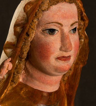 Zdjęcie nr 11: Rzeźba drążona, ustawiona na niewielkim, wielobocznym postumencie. Stojąca Maria ukazana frontalnie, w silnym kontrapoście z esowato wygiętym ciałem. Na lewej ręce podtrzymuje siedzące Dzieciątko, w prawej ręce ugiętej w łokciu i wyciągniętej do przodu najpewniej znajdowało się kiedyś berło.  Maria została ukazana jako młoda kobieta o owalnej twarzy, dużych, migdałowatych oczach, długim, prostym nosie i wyraźnie zaakcentowanych, dużych ustach. Karnacja jasna z rumieńcem zaznaczonym na policzkach i podbródku. Długie, jasne i falowane włosy częściowo zakryte chustą, symetrycznie okalają twarz postaci, spadając na jej ramiona. Maria jest ubrana w długą, sięgającą ziemi suknię, spod której widoczne są ostro zakończone czubki złotych butów oraz obszerny, złoty płaszcz tworzący ostro załamujące się fałdy na wysokości kolan Marii i formujący kaskadę wzdłuż prawego boku postaci. Na głowie Marii znajduje się biała chusta ze złota lamówką. Dzieciątko Jezus ukazane w pozycji siedzącej z lewą nogą wysuniętą wyraźnie do przodu. Prawą ręką wykonuje gest błogosławieństwa, w lewej trzyma niewielkie jabłko. Chłopiec o okrągłej, pulchnej twarzy i  krótkich, falowanych włosach spogląda przed siebie. Ubrany jest w wąską, suknię o długich rękawach, wykończoną przy dekolcie złotą lamówką. Spod sukni widoczne są bose stopy postaci. Karnacje postaci i część szat polichromowane, suknia Marii srebrzona, płaszcz i buty złocone.