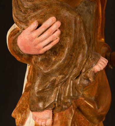 Zdjęcie nr 9: Rzeźba drążona, ustawiona na niewielkim, wielobocznym postumencie. Stojąca Maria ukazana frontalnie, w silnym kontrapoście z esowato wygiętym ciałem. Na lewej ręce podtrzymuje siedzące Dzieciątko, w prawej ręce ugiętej w łokciu i wyciągniętej do przodu najpewniej znajdowało się kiedyś berło.  Maria została ukazana jako młoda kobieta o owalnej twarzy, dużych, migdałowatych oczach, długim, prostym nosie i wyraźnie zaakcentowanych, dużych ustach. Karnacja jasna z rumieńcem zaznaczonym na policzkach i podbródku. Długie, jasne i falowane włosy częściowo zakryte chustą, symetrycznie okalają twarz postaci, spadając na jej ramiona. Maria jest ubrana w długą, sięgającą ziemi suknię, spod której widoczne są ostro zakończone czubki złotych butów oraz obszerny, złoty płaszcz tworzący ostro załamujące się fałdy na wysokości kolan Marii i formujący kaskadę wzdłuż prawego boku postaci. Na głowie Marii znajduje się biała chusta ze złota lamówką. Dzieciątko Jezus ukazane w pozycji siedzącej z lewą nogą wysuniętą wyraźnie do przodu. Prawą ręką wykonuje gest błogosławieństwa, w lewej trzyma niewielkie jabłko. Chłopiec o okrągłej, pulchnej twarzy i  krótkich, falowanych włosach spogląda przed siebie. Ubrany jest w wąską, suknię o długich rękawach, wykończoną przy dekolcie złotą lamówką. Spod sukni widoczne są bose stopy postaci. Karnacje postaci i część szat polichromowane, suknia Marii srebrzona, płaszcz i buty złocone.