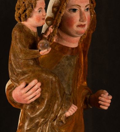 Zdjęcie nr 4: Rzeźba drążona, ustawiona na niewielkim, wielobocznym postumencie. Stojąca Maria ukazana frontalnie, w silnym kontrapoście z esowato wygiętym ciałem. Na lewej ręce podtrzymuje siedzące Dzieciątko, w prawej ręce ugiętej w łokciu i wyciągniętej do przodu najpewniej znajdowało się kiedyś berło.  Maria została ukazana jako młoda kobieta o owalnej twarzy, dużych, migdałowatych oczach, długim, prostym nosie i wyraźnie zaakcentowanych, dużych ustach. Karnacja jasna z rumieńcem zaznaczonym na policzkach i podbródku. Długie, jasne i falowane włosy częściowo zakryte chustą, symetrycznie okalają twarz postaci, spadając na jej ramiona. Maria jest ubrana w długą, sięgającą ziemi suknię, spod której widoczne są ostro zakończone czubki złotych butów oraz obszerny, złoty płaszcz tworzący ostro załamujące się fałdy na wysokości kolan Marii i formujący kaskadę wzdłuż prawego boku postaci. Na głowie Marii znajduje się biała chusta ze złota lamówką. Dzieciątko Jezus ukazane w pozycji siedzącej z lewą nogą wysuniętą wyraźnie do przodu. Prawą ręką wykonuje gest błogosławieństwa, w lewej trzyma niewielkie jabłko. Chłopiec o okrągłej, pulchnej twarzy i  krótkich, falowanych włosach spogląda przed siebie. Ubrany jest w wąską, suknię o długich rękawach, wykończoną przy dekolcie złotą lamówką. Spod sukni widoczne są bose stopy postaci. Karnacje postaci i część szat polichromowane, suknia Marii srebrzona, płaszcz i buty złocone.