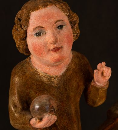 Zdjęcie nr 14: Rzeźba drążona, ustawiona na niewielkim, wielobocznym postumencie. Stojąca Maria ukazana frontalnie, w silnym kontrapoście z esowato wygiętym ciałem. Na lewej ręce podtrzymuje siedzące Dzieciątko, w prawej ręce ugiętej w łokciu i wyciągniętej do przodu najpewniej znajdowało się kiedyś berło.  Maria została ukazana jako młoda kobieta o owalnej twarzy, dużych, migdałowatych oczach, długim, prostym nosie i wyraźnie zaakcentowanych, dużych ustach. Karnacja jasna z rumieńcem zaznaczonym na policzkach i podbródku. Długie, jasne i falowane włosy częściowo zakryte chustą, symetrycznie okalają twarz postaci, spadając na jej ramiona. Maria jest ubrana w długą, sięgającą ziemi suknię, spod której widoczne są ostro zakończone czubki złotych butów oraz obszerny, złoty płaszcz tworzący ostro załamujące się fałdy na wysokości kolan Marii i formujący kaskadę wzdłuż prawego boku postaci. Na głowie Marii znajduje się biała chusta ze złota lamówką. Dzieciątko Jezus ukazane w pozycji siedzącej z lewą nogą wysuniętą wyraźnie do przodu. Prawą ręką wykonuje gest błogosławieństwa, w lewej trzyma niewielkie jabłko. Chłopiec o okrągłej, pulchnej twarzy i  krótkich, falowanych włosach spogląda przed siebie. Ubrany jest w wąską, suknię o długich rękawach, wykończoną przy dekolcie złotą lamówką. Spod sukni widoczne są bose stopy postaci. Karnacje postaci i część szat polichromowane, suknia Marii srebrzona, płaszcz i buty złocone.