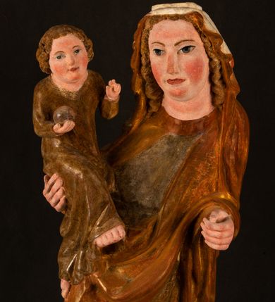 Zdjęcie nr 23: Rzeźba drążona, ustawiona na niewielkim, wielobocznym postumencie. Stojąca Maria ukazana frontalnie, w silnym kontrapoście z esowato wygiętym ciałem. Na lewej ręce podtrzymuje siedzące Dzieciątko, w prawej ręce ugiętej w łokciu i wyciągniętej do przodu najpewniej znajdowało się kiedyś berło.  Maria została ukazana jako młoda kobieta o owalnej twarzy, dużych, migdałowatych oczach, długim, prostym nosie i wyraźnie zaakcentowanych, dużych ustach. Karnacja jasna z rumieńcem zaznaczonym na policzkach i podbródku. Długie, jasne i falowane włosy częściowo zakryte chustą, symetrycznie okalają twarz postaci, spadając na jej ramiona. Maria jest ubrana w długą, sięgającą ziemi suknię, spod której widoczne są ostro zakończone czubki złotych butów oraz obszerny, złoty płaszcz tworzący ostro załamujące się fałdy na wysokości kolan Marii i formujący kaskadę wzdłuż prawego boku postaci. Na głowie Marii znajduje się biała chusta ze złota lamówką. Dzieciątko Jezus ukazane w pozycji siedzącej z lewą nogą wysuniętą wyraźnie do przodu. Prawą ręką wykonuje gest błogosławieństwa, w lewej trzyma niewielkie jabłko. Chłopiec o okrągłej, pulchnej twarzy i  krótkich, falowanych włosach spogląda przed siebie. Ubrany jest w wąską, suknię o długich rękawach, wykończoną przy dekolcie złotą lamówką. Spod sukni widoczne są bose stopy postaci. Karnacje postaci i część szat polichromowane, suknia Marii srebrzona, płaszcz i buty złocone.