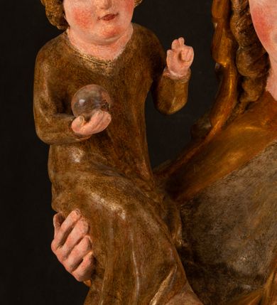 Zdjęcie nr 8: Rzeźba drążona, ustawiona na niewielkim, wielobocznym postumencie. Stojąca Maria ukazana frontalnie, w silnym kontrapoście z esowato wygiętym ciałem. Na lewej ręce podtrzymuje siedzące Dzieciątko, w prawej ręce ugiętej w łokciu i wyciągniętej do przodu najpewniej znajdowało się kiedyś berło.  Maria została ukazana jako młoda kobieta o owalnej twarzy, dużych, migdałowatych oczach, długim, prostym nosie i wyraźnie zaakcentowanych, dużych ustach. Karnacja jasna z rumieńcem zaznaczonym na policzkach i podbródku. Długie, jasne i falowane włosy częściowo zakryte chustą, symetrycznie okalają twarz postaci, spadając na jej ramiona. Maria jest ubrana w długą, sięgającą ziemi suknię, spod której widoczne są ostro zakończone czubki złotych butów oraz obszerny, złoty płaszcz tworzący ostro załamujące się fałdy na wysokości kolan Marii i formujący kaskadę wzdłuż prawego boku postaci. Na głowie Marii znajduje się biała chusta ze złota lamówką. Dzieciątko Jezus ukazane w pozycji siedzącej z lewą nogą wysuniętą wyraźnie do przodu. Prawą ręką wykonuje gest błogosławieństwa, w lewej trzyma niewielkie jabłko. Chłopiec o okrągłej, pulchnej twarzy i  krótkich, falowanych włosach spogląda przed siebie. Ubrany jest w wąską, suknię o długich rękawach, wykończoną przy dekolcie złotą lamówką. Spod sukni widoczne są bose stopy postaci. Karnacje postaci i część szat polichromowane, suknia Marii srebrzona, płaszcz i buty złocone.