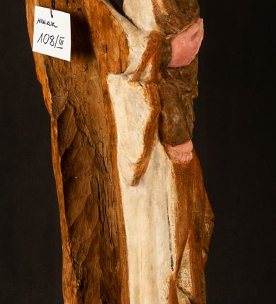 Zdjęcie nr 20: Rzeźba drążona, ustawiona na niewielkim, wielobocznym postumencie. Stojąca Maria ukazana frontalnie, w silnym kontrapoście z esowato wygiętym ciałem. Na lewej ręce podtrzymuje siedzące Dzieciątko, w prawej ręce ugiętej w łokciu i wyciągniętej do przodu najpewniej znajdowało się kiedyś berło.  Maria została ukazana jako młoda kobieta o owalnej twarzy, dużych, migdałowatych oczach, długim, prostym nosie i wyraźnie zaakcentowanych, dużych ustach. Karnacja jasna z rumieńcem zaznaczonym na policzkach i podbródku. Długie, jasne i falowane włosy częściowo zakryte chustą, symetrycznie okalają twarz postaci, spadając na jej ramiona. Maria jest ubrana w długą, sięgającą ziemi suknię, spod której widoczne są ostro zakończone czubki złotych butów oraz obszerny, złoty płaszcz tworzący ostro załamujące się fałdy na wysokości kolan Marii i formujący kaskadę wzdłuż prawego boku postaci. Na głowie Marii znajduje się biała chusta ze złota lamówką. Dzieciątko Jezus ukazane w pozycji siedzącej z lewą nogą wysuniętą wyraźnie do przodu. Prawą ręką wykonuje gest błogosławieństwa, w lewej trzyma niewielkie jabłko. Chłopiec o okrągłej, pulchnej twarzy i  krótkich, falowanych włosach spogląda przed siebie. Ubrany jest w wąską, suknię o długich rękawach, wykończoną przy dekolcie złotą lamówką. Spod sukni widoczne są bose stopy postaci. Karnacje postaci i część szat polichromowane, suknia Marii srebrzona, płaszcz i buty złocone.