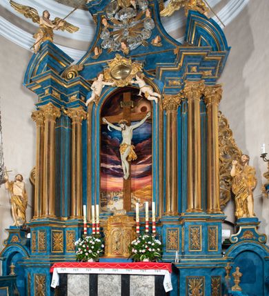 Zdjęcie nr 27: Ołtarz architektoniczny, jednoosiowy, jednokondygnacyjny ze zwieńczeniem. Na zdwojonych cokołach usytuowanych ukośnie względem osi po trzy kompozytowe kolumny opinające filary o gładkich trzonach podtrzymujących wyłamujące się nad nimi belkowanie z ćwierćkolistymi naczółkami. Na nich siedzące anioły z narzędzami męki Pańskiej. Pośrodku wnęka zamknięta półkoliście i wchodząca w partię belkowania z figurą Ukrzyżowanego. Nad wnęką dwa nagie putta trzymają kartusz z napisem: &quot;DZIEŁO / NIE MAJĄC PRZYKŁADU / ŁASKA NIE ZASŁUŻONA / DAR NIE OCENIONY / MIŁOŚĆ BEZ GRANIC&quot;.  Zwieńczenie ujęte spływami wolutowymi, mocno zwężające się ku górze, zakończone gzymsem w formie łuku nadwieszonego. W polu owalny prześwit z glorią promienistą w chmurach z figurą Boga Ojca, puttami siedzącymi na gzymsie i główkami aniołów wśród chmur. Po bokach ołtarza dwie bramki z cokołami ujętymi spływami wolutowymi, na których figury św. Jana Chrzciciela i św. Jana Ewangelisty. Dekoracja ornamentalna w postaci rozłożystych liści na cokołach oraz liściastych, ażurowych uszy ujmujących nastawę. 