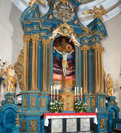 Zdjęcie nr 60: Ołtarz architektoniczny, jednoosiowy, jednokondygnacyjny ze zwieńczeniem. Na zdwojonych cokołach usytuowanych ukośnie względem osi po trzy kompozytowe kolumny opinające filary o gładkich trzonach podtrzymujących wyłamujące się nad nimi belkowanie z ćwierćkolistymi naczółkami. Na nich siedzące anioły z narzędzami męki Pańskiej. Pośrodku wnęka zamknięta półkoliście i wchodząca w partię belkowania z figurą Ukrzyżowanego. Nad wnęką dwa nagie putta trzymają kartusz z napisem: "DZIEŁO / NIE MAJĄC PRZYKŁADU / ŁASKA NIE ZASŁUŻONA / DAR NIE OCENIONY / MIŁOŚĆ BEZ GRANIC".  Zwieńczenie ujęte spływami wolutowymi, mocno zwężające się ku górze, zakończone gzymsem w formie łuku nadwieszonego. W polu owalny prześwit z glorią promienistą w chmurach z figurą Boga Ojca, puttami siedzącymi na gzymsie i główkami aniołów wśród chmur. Po bokach ołtarza dwie bramki z cokołami ujętymi spływami wolutowymi, na których figury św. Jana Chrzciciela i św. Jana Ewangelisty. Dekoracja ornamentalna w postaci rozłożystych liści na cokołach oraz liściastych, ażurowych uszy ujmujących nastawę. 