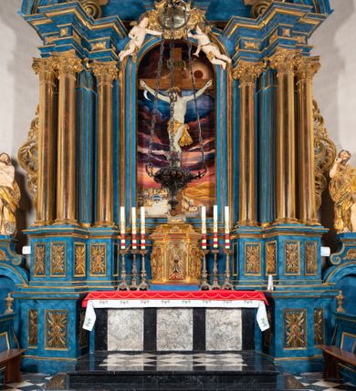 Zdjęcie nr 81: Ołtarz architektoniczny, jednoosiowy, jednokondygnacyjny ze zwieńczeniem. Na zdwojonych cokołach usytuowanych ukośnie względem osi po trzy kompozytowe kolumny opinające filary o gładkich trzonach podtrzymujących wyłamujące się nad nimi belkowanie z ćwierćkolistymi naczółkami. Na nich siedzące anioły z narzędzami męki Pańskiej. Pośrodku wnęka zamknięta półkoliście i wchodząca w partię belkowania z figurą Ukrzyżowanego. Nad wnęką dwa nagie putta trzymają kartusz z napisem: "DZIEŁO / NIE MAJĄC PRZYKŁADU / ŁASKA NIE ZASŁUŻONA / DAR NIE OCENIONY / MIŁOŚĆ BEZ GRANIC".  Zwieńczenie ujęte spływami wolutowymi, mocno zwężające się ku górze, zakończone gzymsem w formie łuku nadwieszonego. W polu owalny prześwit z glorią promienistą w chmurach z figurą Boga Ojca, puttami siedzącymi na gzymsie i główkami aniołów wśród chmur. Po bokach ołtarza dwie bramki z cokołami ujętymi spływami wolutowymi, na których figury św. Jana Chrzciciela i św. Jana Ewangelisty. Dekoracja ornamentalna w postaci rozłożystych liści na cokołach oraz liściastych, ażurowych uszy ujmujących nastawę. 