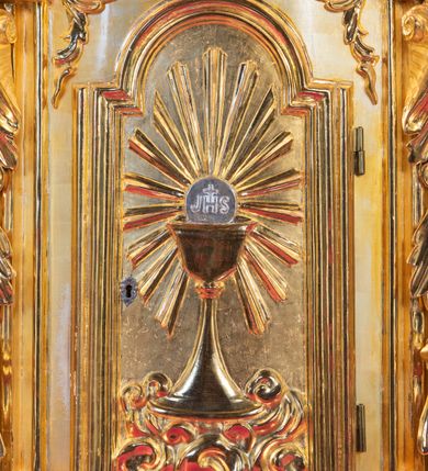 Zdjęcie nr 9: Ołtarz architektoniczny, jednoosiowy, jednokondygnacyjny ze zwieńczeniem. Na zdwojonych cokołach usytuowanych ukośnie względem osi po trzy kompozytowe kolumny opinające filary o gładkich trzonach podtrzymujących wyłamujące się nad nimi belkowanie z ćwierćkolistymi naczółkami. Na nich siedzące anioły z narzędzami męki Pańskiej. Pośrodku wnęka zamknięta półkoliście i wchodząca w partię belkowania z figurą Ukrzyżowanego. Nad wnęką dwa nagie putta trzymają kartusz z napisem: "DZIEŁO / NIE MAJĄC PRZYKŁADU / ŁASKA NIE ZASŁUŻONA / DAR NIE OCENIONY / MIŁOŚĆ BEZ GRANIC".  Zwieńczenie ujęte spływami wolutowymi, mocno zwężające się ku górze, zakończone gzymsem w formie łuku nadwieszonego. W polu owalny prześwit z glorią promienistą w chmurach z figurą Boga Ojca, puttami siedzącymi na gzymsie i główkami aniołów wśród chmur. Po bokach ołtarza dwie bramki z cokołami ujętymi spływami wolutowymi, na których figury św. Jana Chrzciciela i św. Jana Ewangelisty. Dekoracja ornamentalna w postaci rozłożystych liści na cokołach oraz liściastych, ażurowych uszy ujmujących nastawę. 