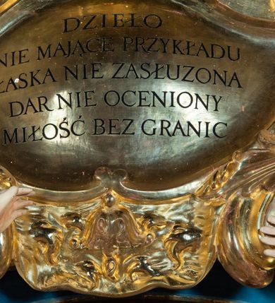 Zdjęcie nr 8: Ołtarz architektoniczny, jednoosiowy, jednokondygnacyjny ze zwieńczeniem. Na zdwojonych cokołach usytuowanych ukośnie względem osi po trzy kompozytowe kolumny opinające filary o gładkich trzonach podtrzymujących wyłamujące się nad nimi belkowanie z ćwierćkolistymi naczółkami. Na nich siedzące anioły z narzędzami męki Pańskiej. Pośrodku wnęka zamknięta półkoliście i wchodząca w partię belkowania z figurą Ukrzyżowanego. Nad wnęką dwa nagie putta trzymają kartusz z napisem: &quot;DZIEŁO / NIE MAJĄC PRZYKŁADU / ŁASKA NIE ZASŁUŻONA / DAR NIE OCENIONY / MIŁOŚĆ BEZ GRANIC&quot;.  Zwieńczenie ujęte spływami wolutowymi, mocno zwężające się ku górze, zakończone gzymsem w formie łuku nadwieszonego. W polu owalny prześwit z glorią promienistą w chmurach z figurą Boga Ojca, puttami siedzącymi na gzymsie i główkami aniołów wśród chmur. Po bokach ołtarza dwie bramki z cokołami ujętymi spływami wolutowymi, na których figury św. Jana Chrzciciela i św. Jana Ewangelisty. Dekoracja ornamentalna w postaci rozłożystych liści na cokołach oraz liściastych, ażurowych uszy ujmujących nastawę. 